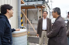 雨水タンクの前で飯塚自治会長さん（中央）と話す。使い方がわからず、タンクのふたが壊されたことがあり、予防でガムテープを貼っているという。